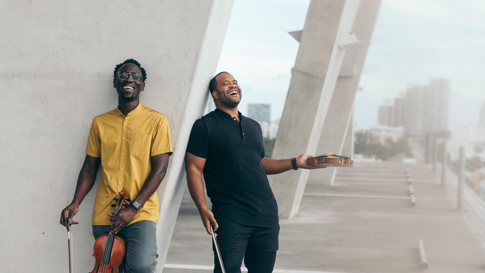 Two men stand against a pillar and laugh while holding violins