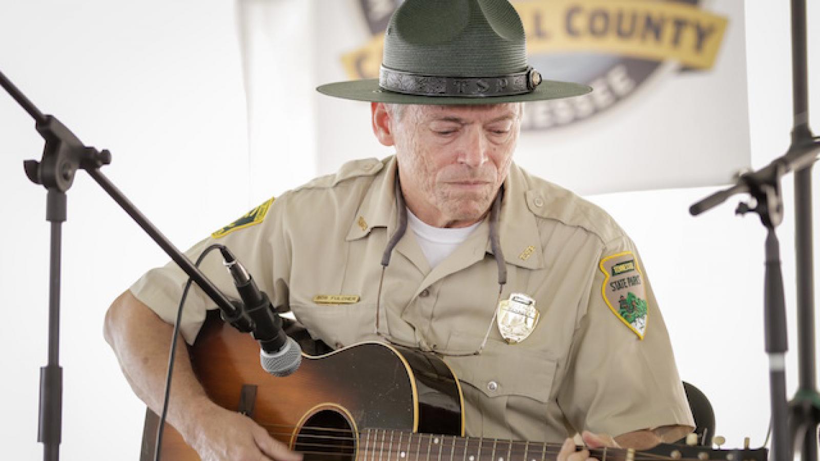 Bob Fulcher in his state parks uniform sits on a chair playing the guitar