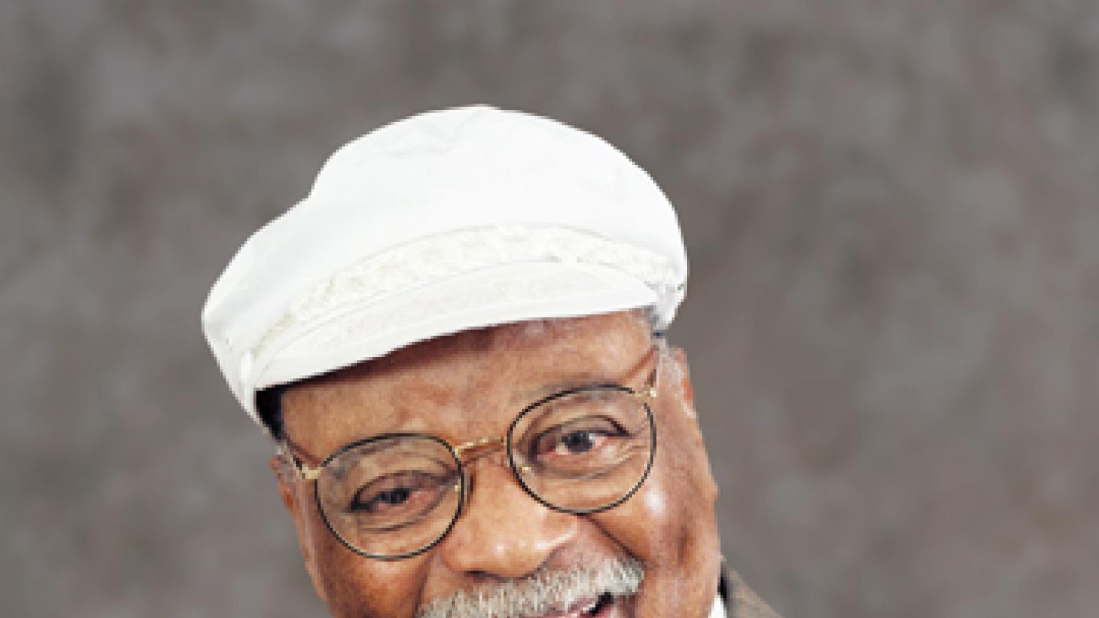 A man in a white hat, with glasses and a mustache smiles, wearing a small trumpet charm on a necklace.