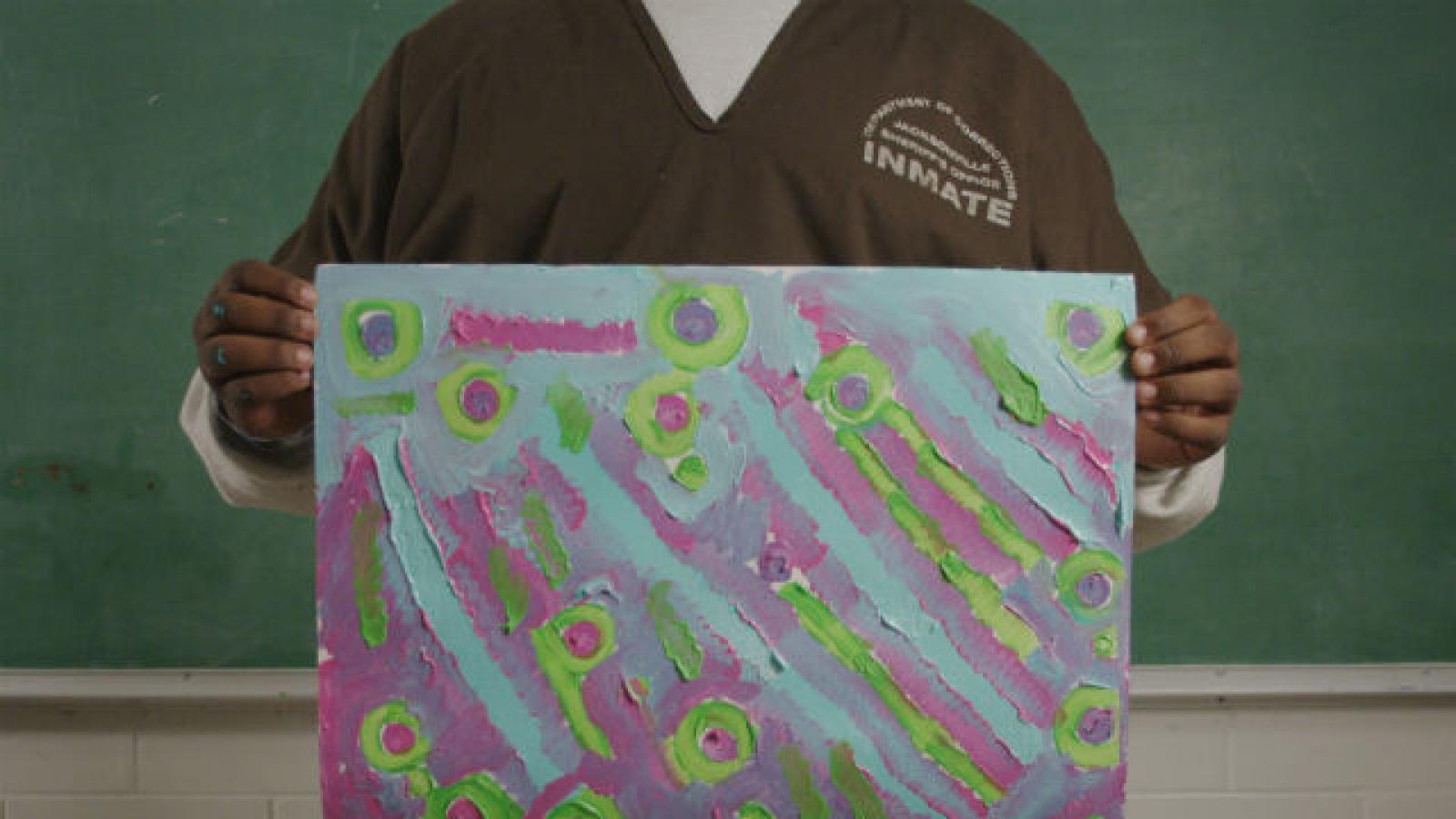 A participant in a juvenile justice arts program A young man in prison garb holds up a brightly colored painting that he made