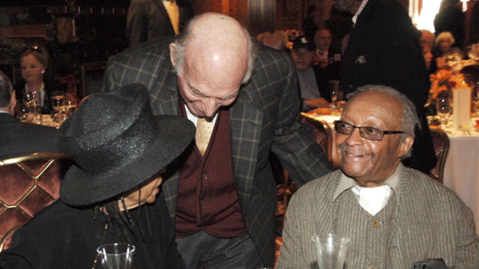 A man and a woman sit at a table at a restaurant with a second man standing between them talking to them.