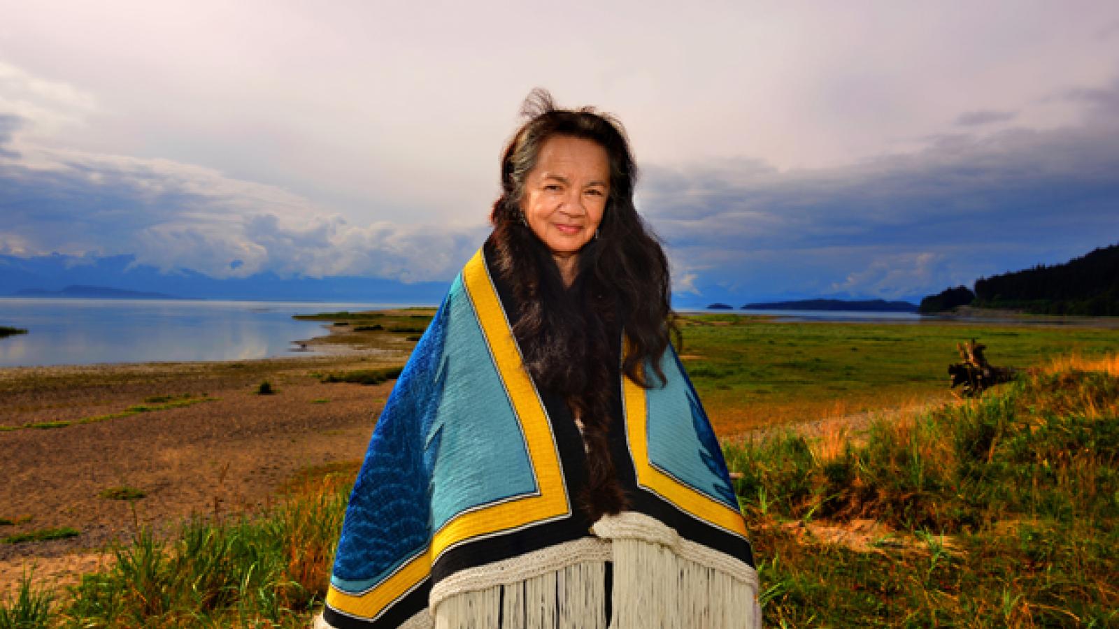 A woman stands on a shore wearing a woven blue and yellow blanket.