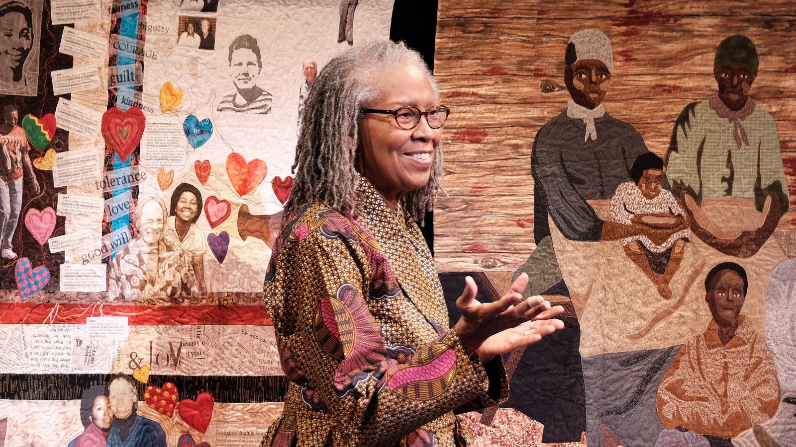 A woman stands smiling with a quilt hanging behind her.