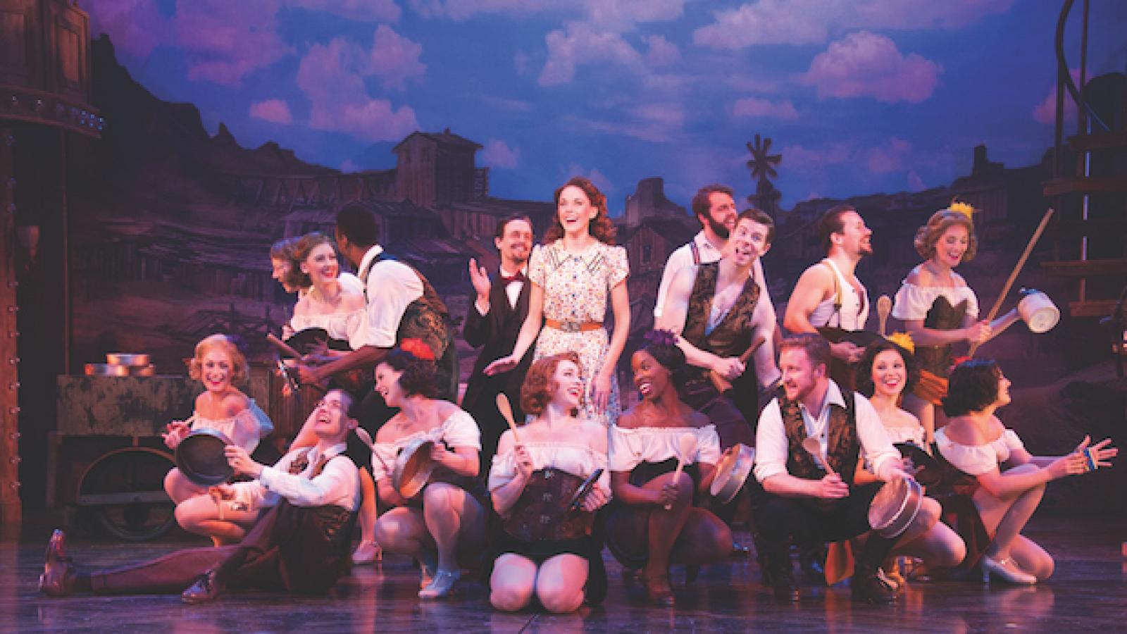 an ensemble of actors posed in a group on a stage holding various pots and pans and other implements