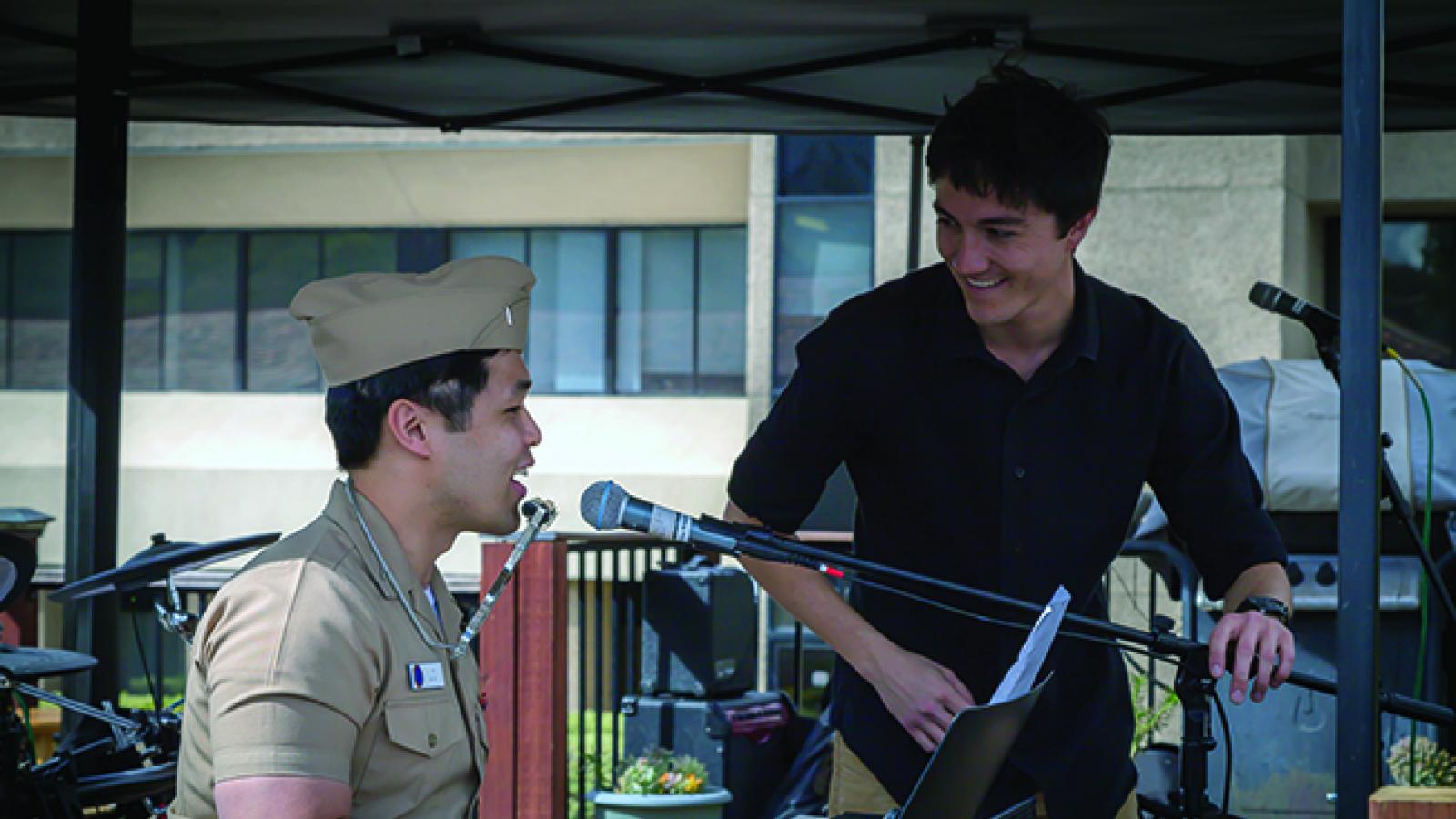 A man in brown Navy uniform plays the piano and wears a harmonica on a neckstrap while another man looks on