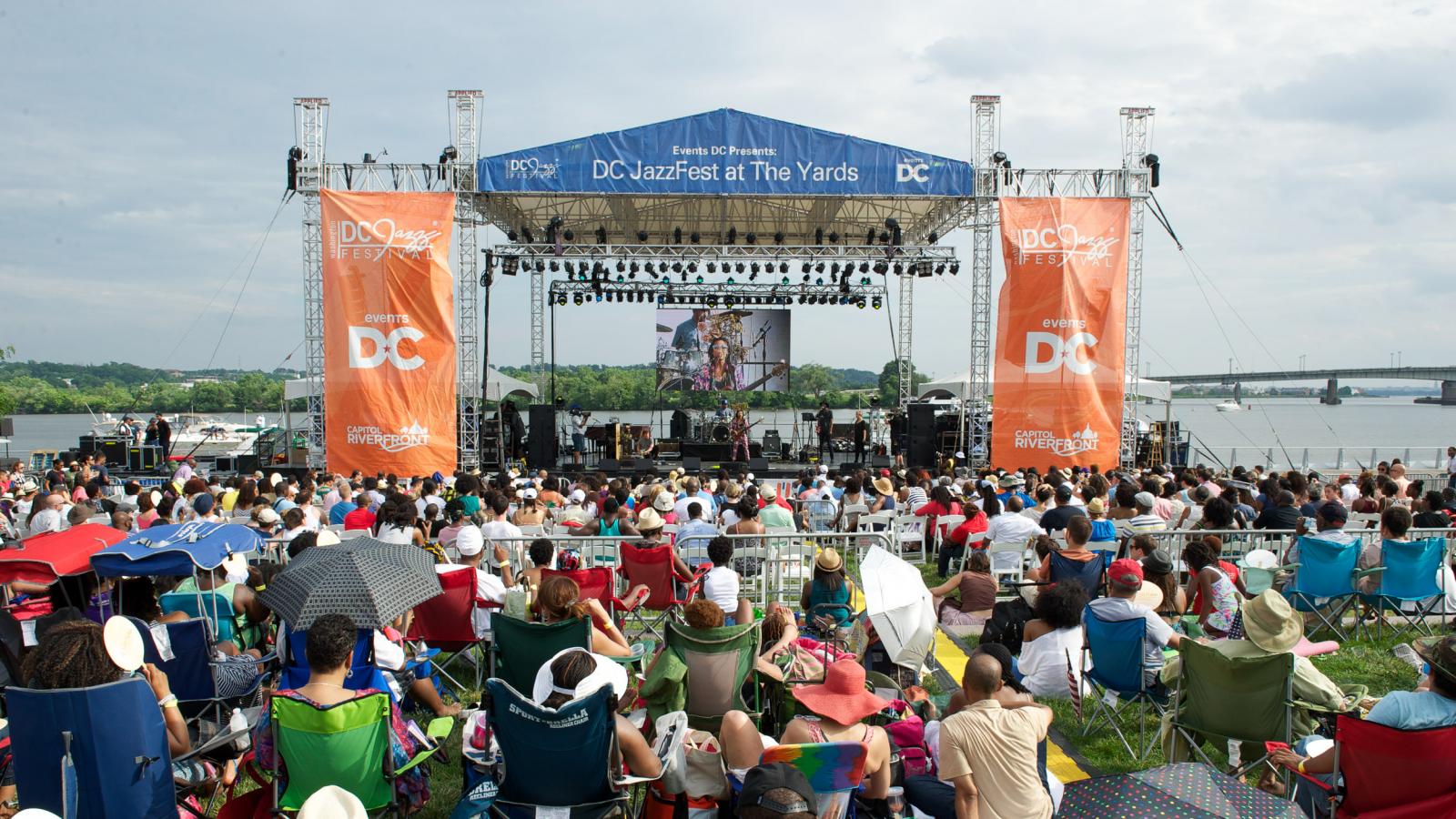 Front view of the outdoor stage with audience members sitting on blankets and folded chairs.