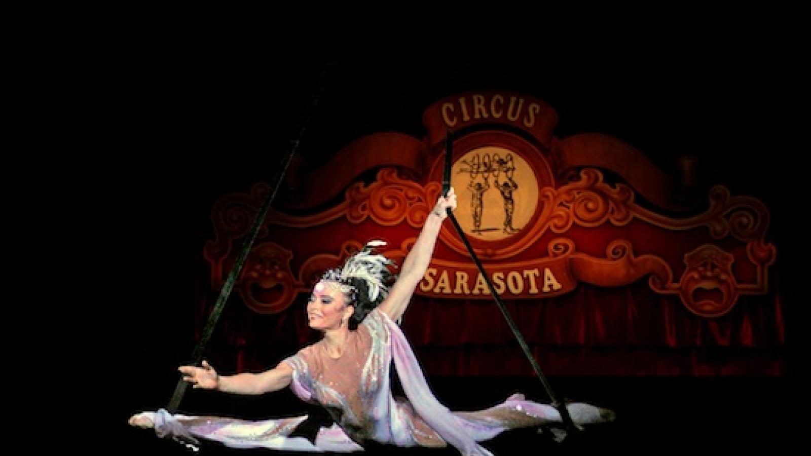 a woman circus aerialist suspended in a split in front of a red circus sarasota sign