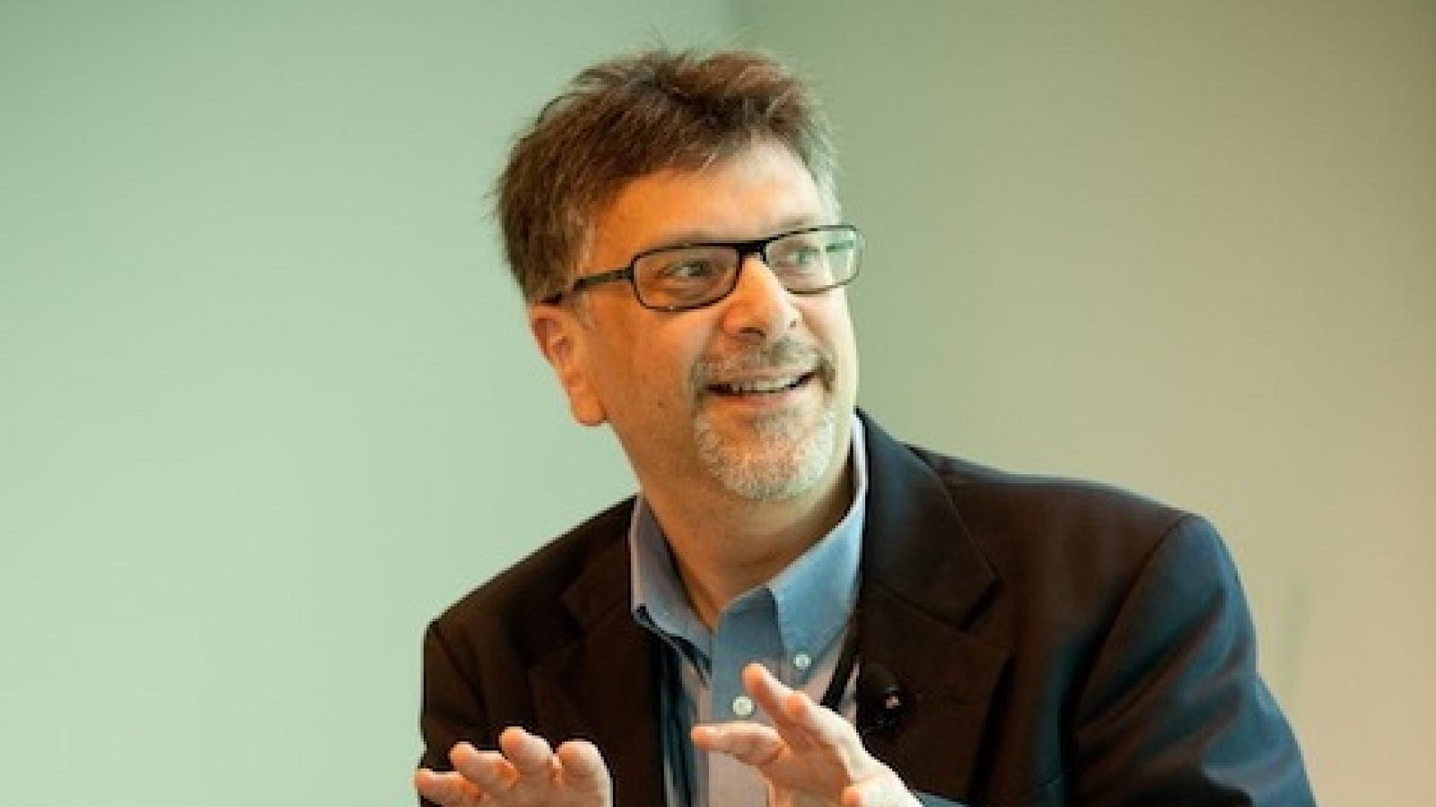 a bearded white man with glasses seated in a chair gesturing with his hands