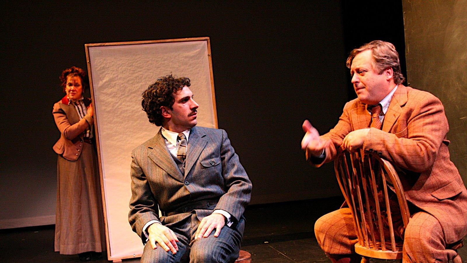 Two men sitting in wooden chairs talking, a woman listening behind a screen. 