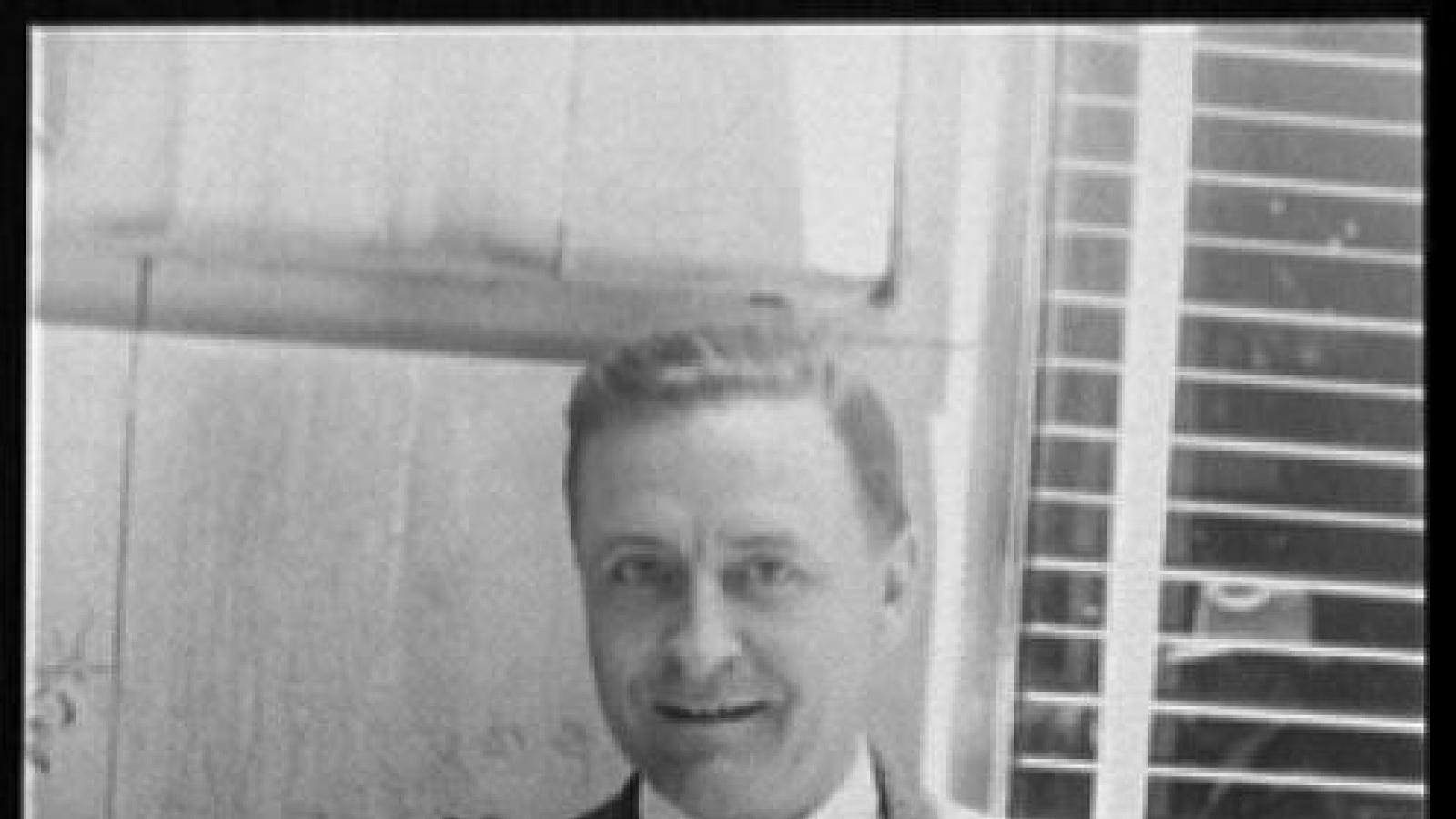 Headshot of F. Scott Fitzgerald. He is standing outside in front of a window and smiling.