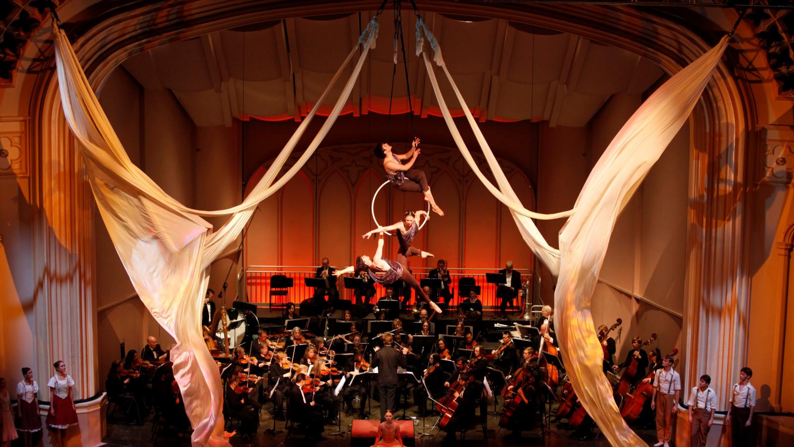 Orchestra on stage performing, while acrobats are also performing on the same stage. 