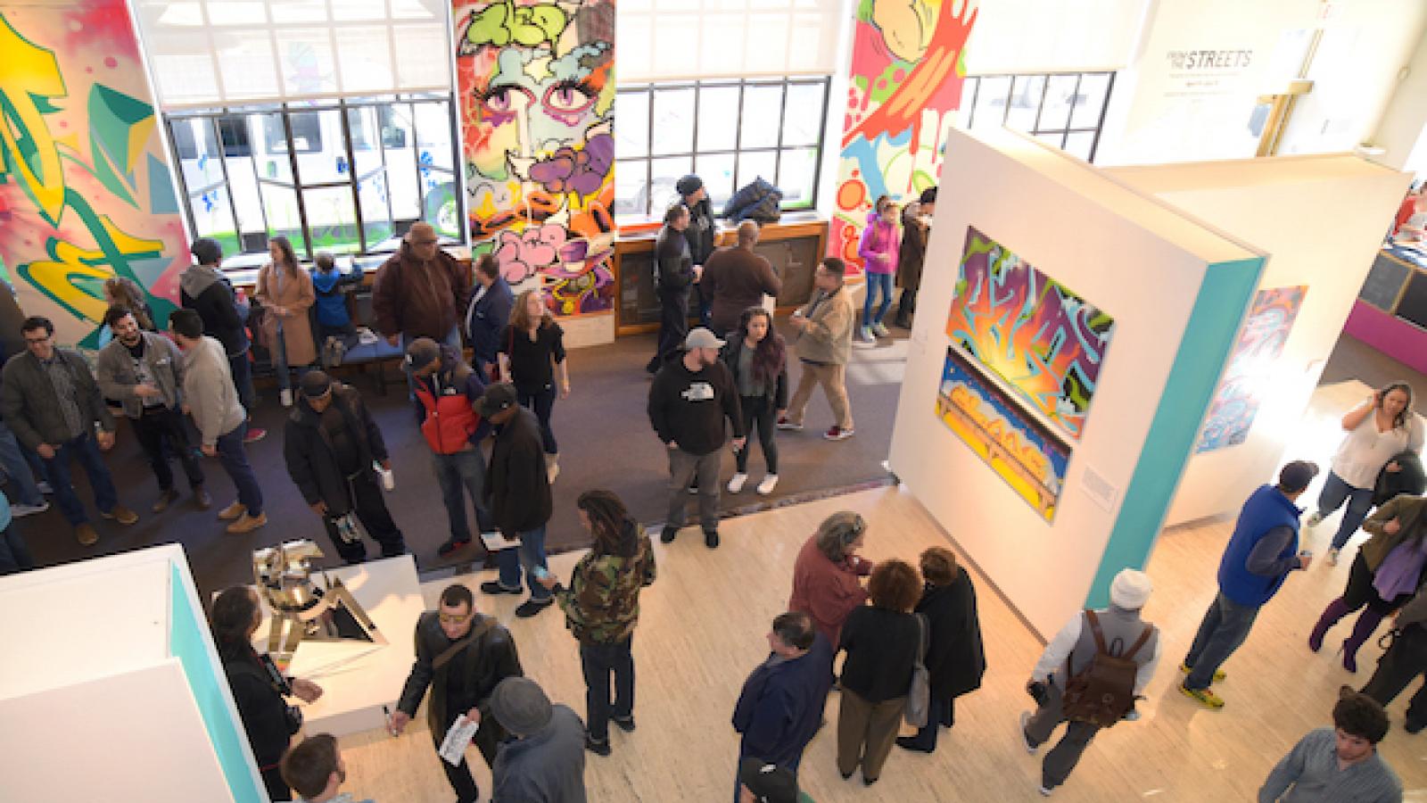 a crowd gathers in an exhibit space with graffiti-inspired artwork is on view