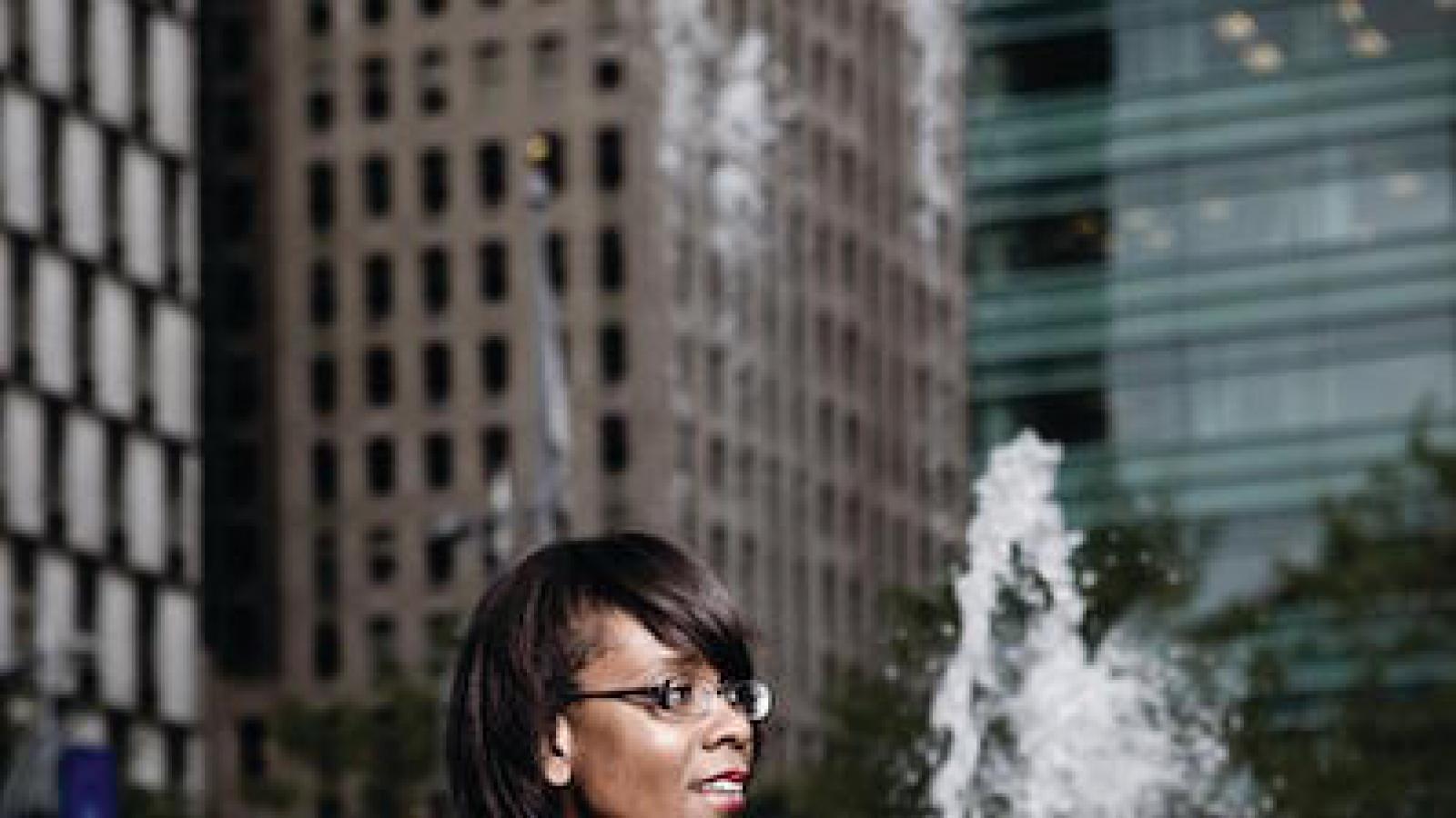 a black woman with glasses in black clothes and a brown leather jacket sitting on the edge of a fountain