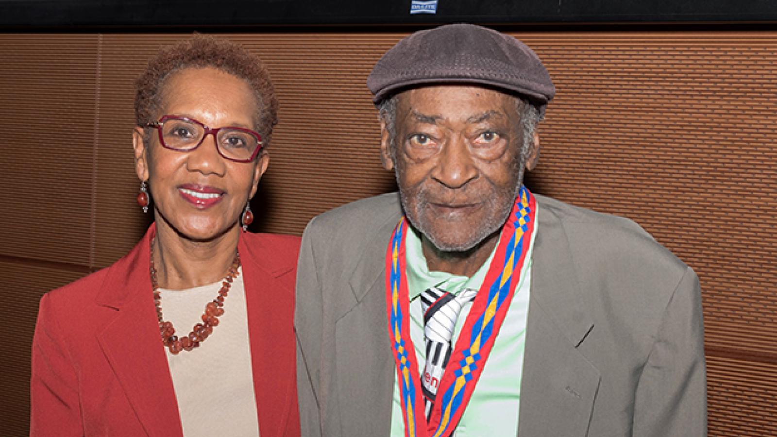 A man in a gray suit wearing a gold medal with a red, blue, and yellow ribbon stands next to a woman in a red suit.