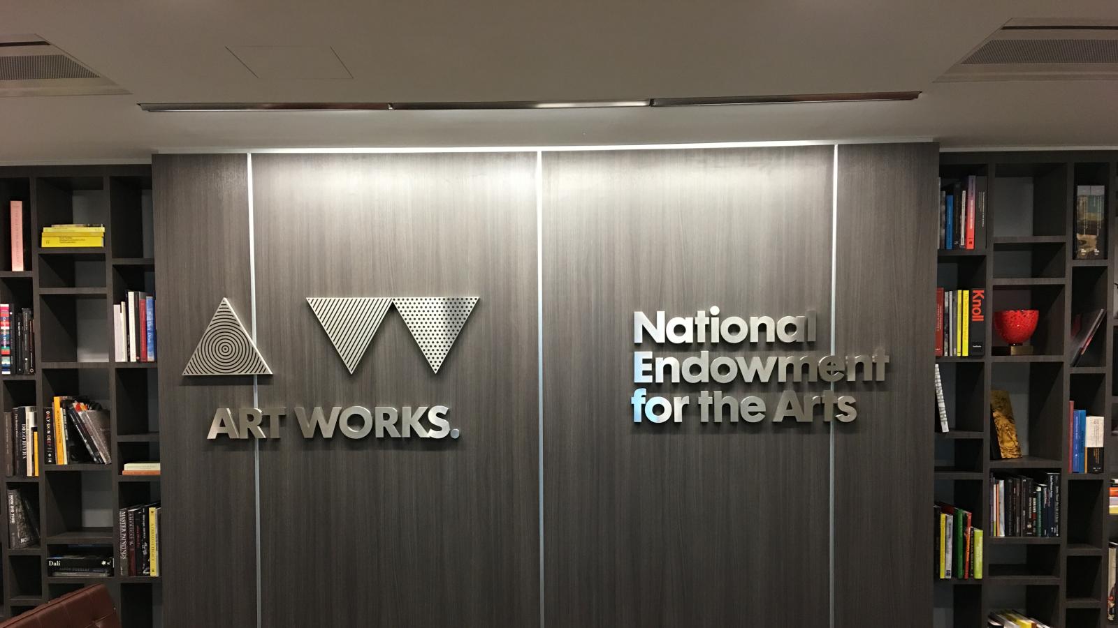 NEA's logo on a gray wall with a bookshelf on each side. 