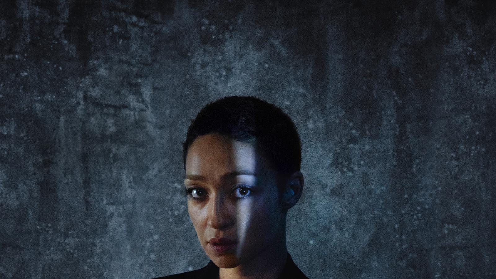 Woman with close-cropped hair in shadows wearing a suit jacket and staring at the camera