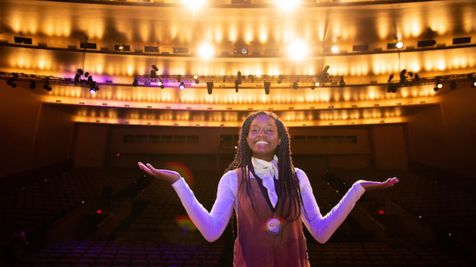 photo of young Black woman with long braids standing on stage with her back to audience and lights blazing behind her