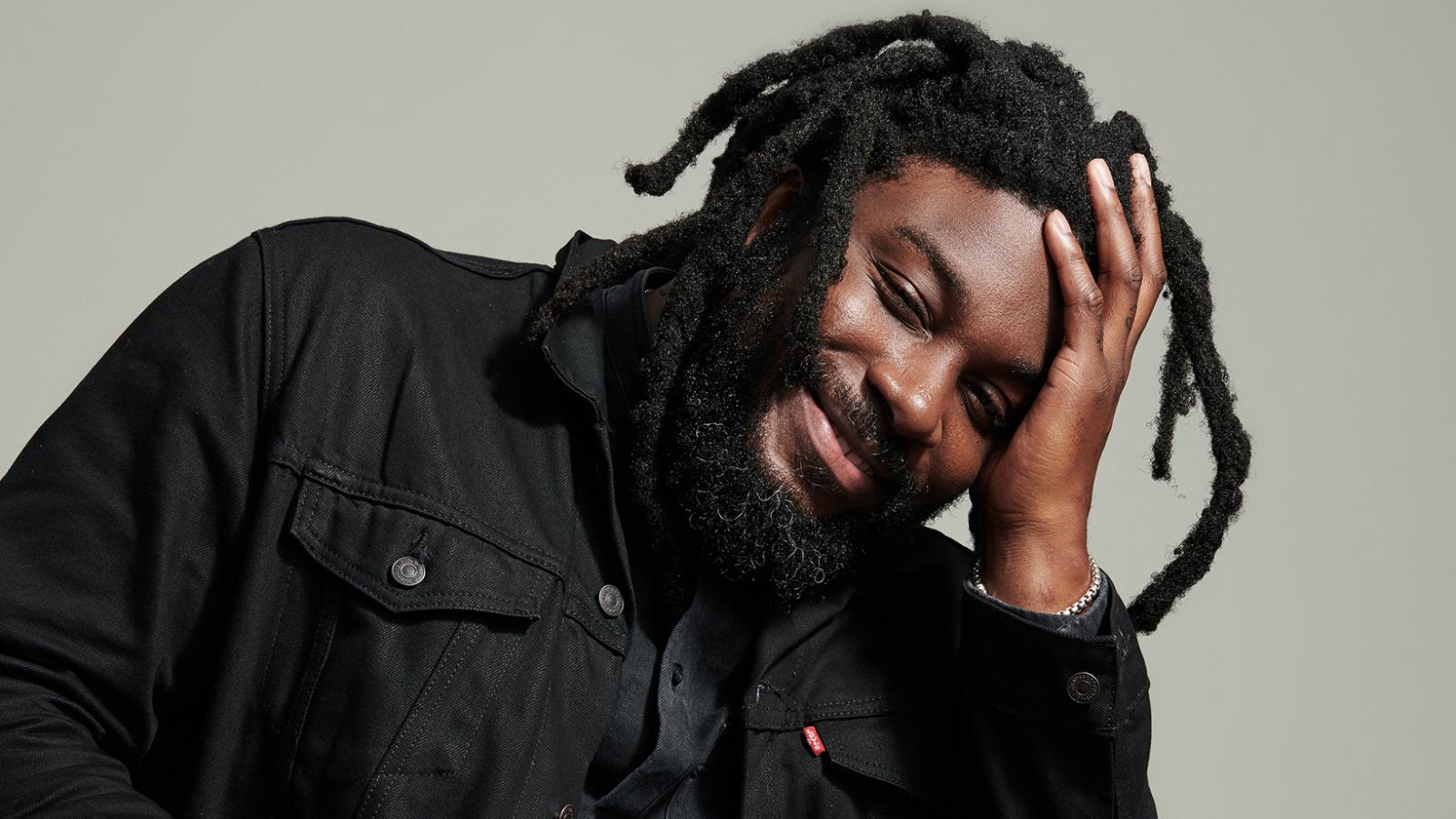 Jason Reynolds Man in black jacket and dreadlocks sits smiling with his head resting on one hand