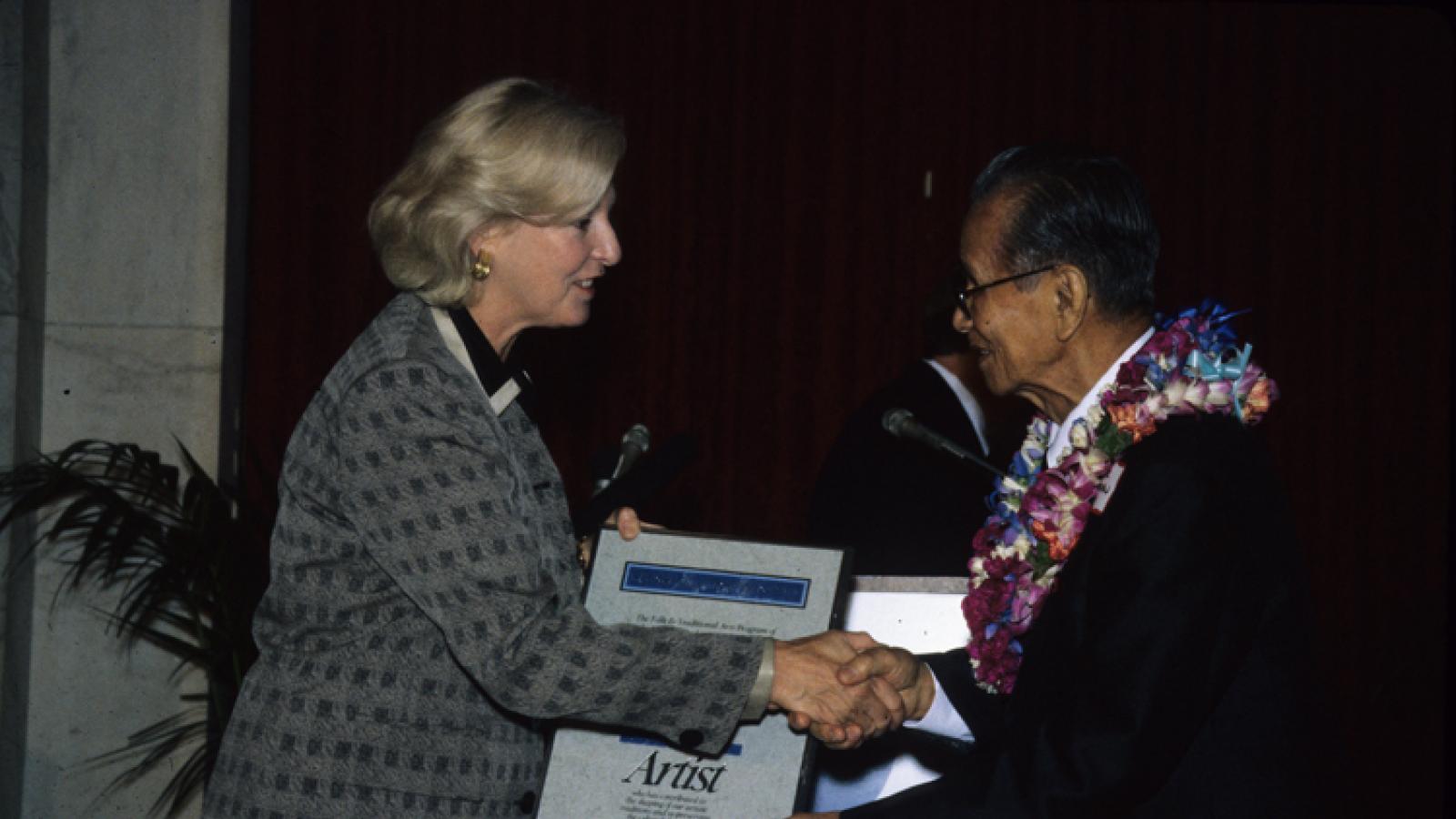 A woman with blond hair presents a large framed certificate to a man in glasses wearing a lei.