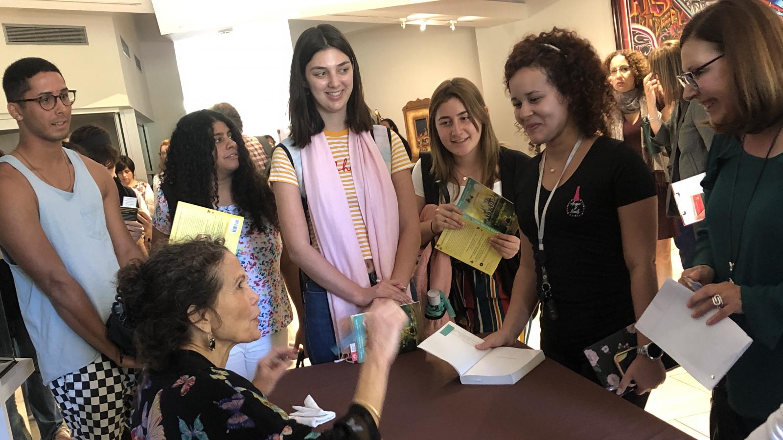 A woman sits behind a desk signing books with a crowd of people around her.