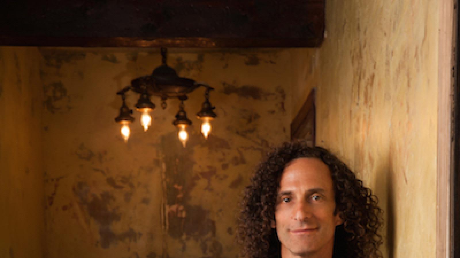 a full-shot of musician Kenny G wearing a dark blue suit and holding a soprano saxophone