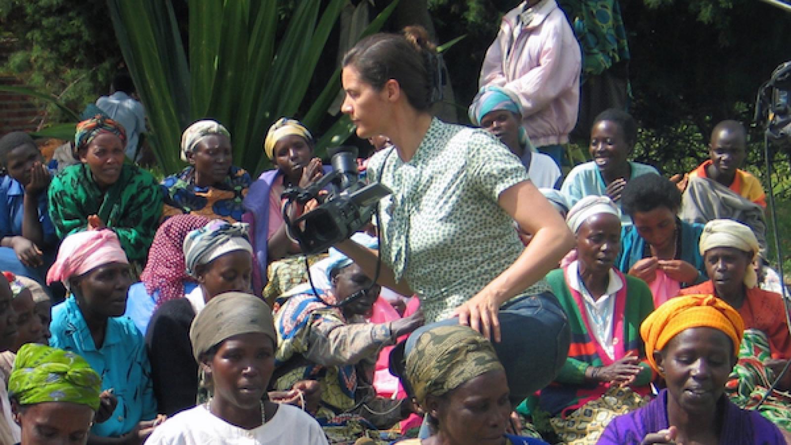  a white woman in Western dress holds a film camera while sitting amidst a crowd of Rwandan women 