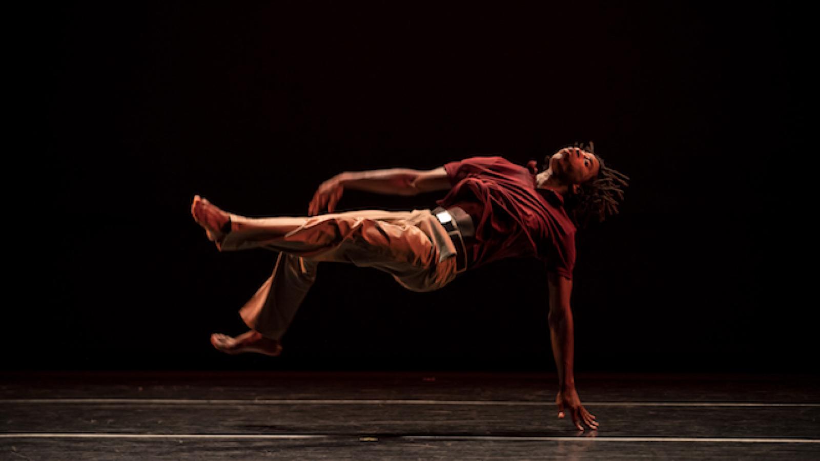 During a dance performance, Cameron McKinney, an African American man with locked hair, seems to float suspended in the air on his back, with just one hand touching the stage.