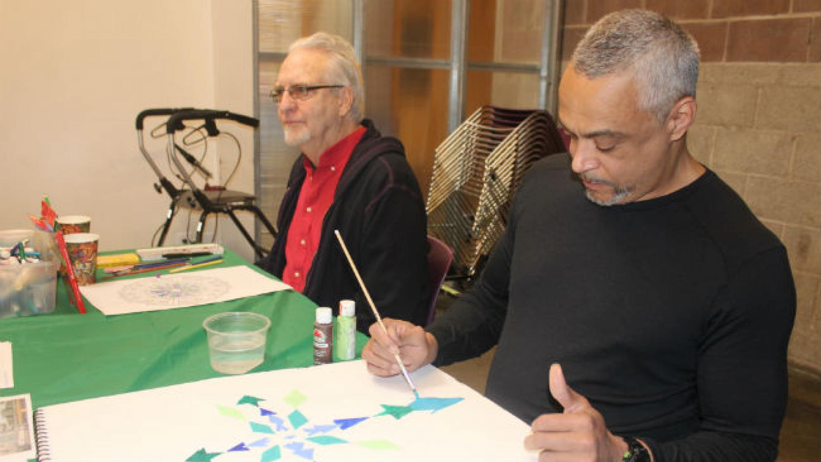 Two older men sit and paint at a table