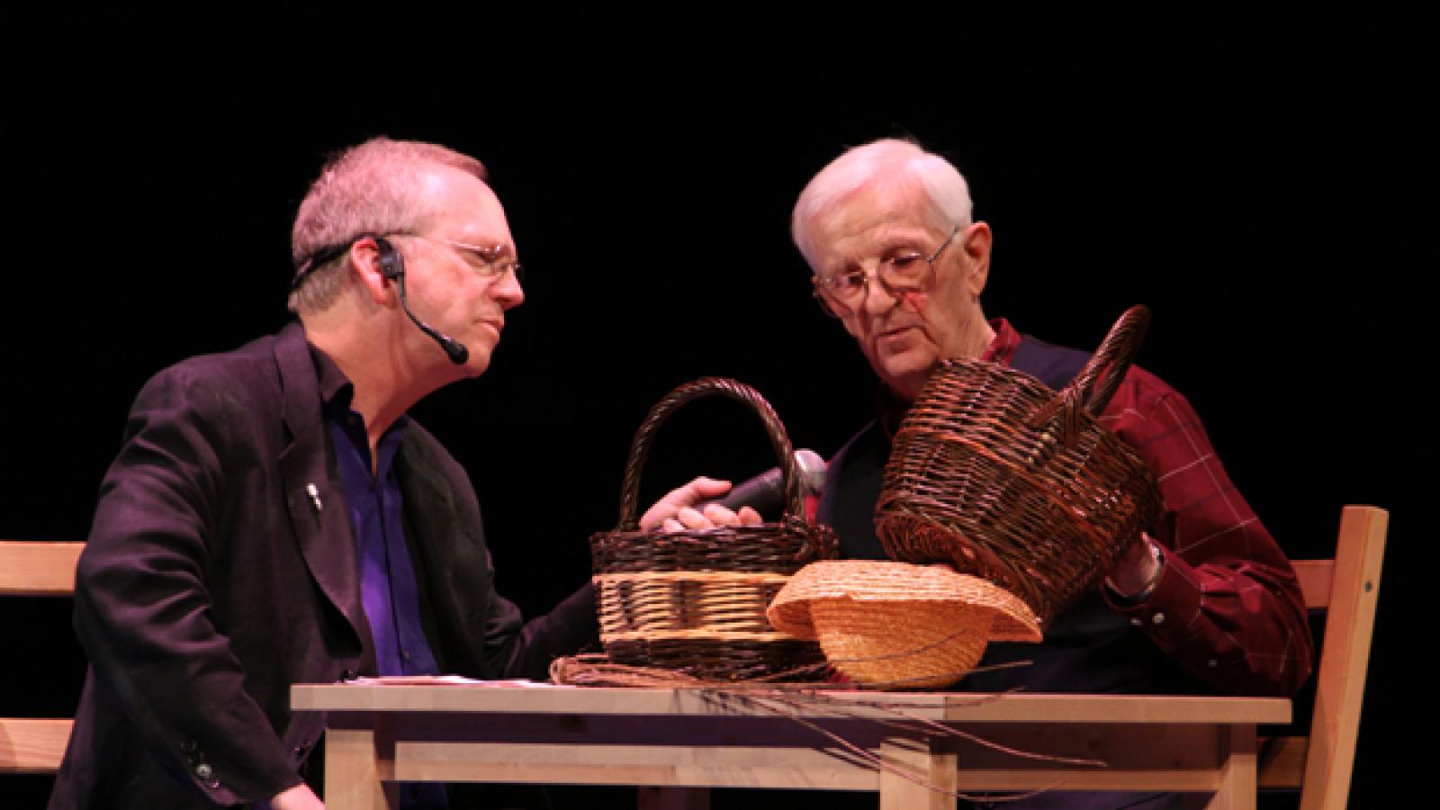 A man in a suit and a man in a red plaid shirt and black vest look at two baskets.