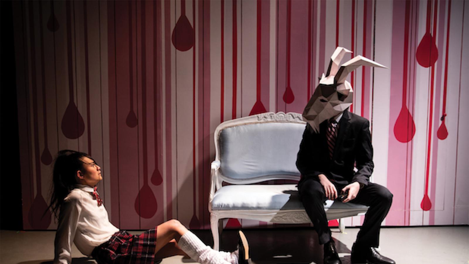 a girl in a school uniform sits on the floor looking up at a person sitting on a couch wearing an animal head