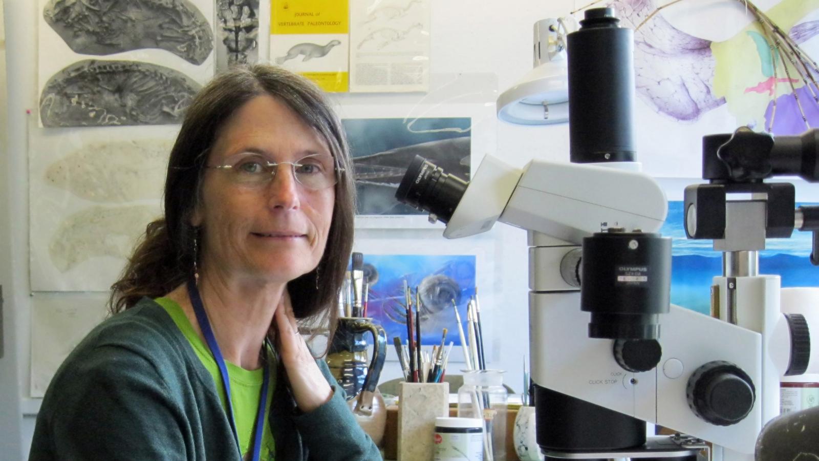 Woman sitting at desk with large microscope while holding pen with art supplies in background