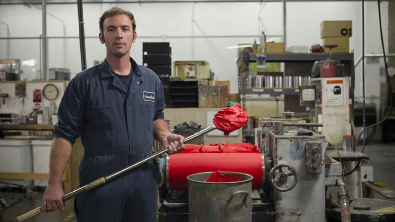 Man standing in front of paint rolling machinery holding a pole with red paint on end. 