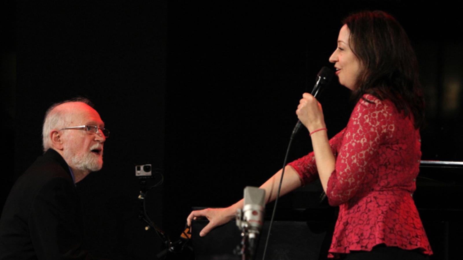 A man performs at the piano with a woman standing next to the piano singing.