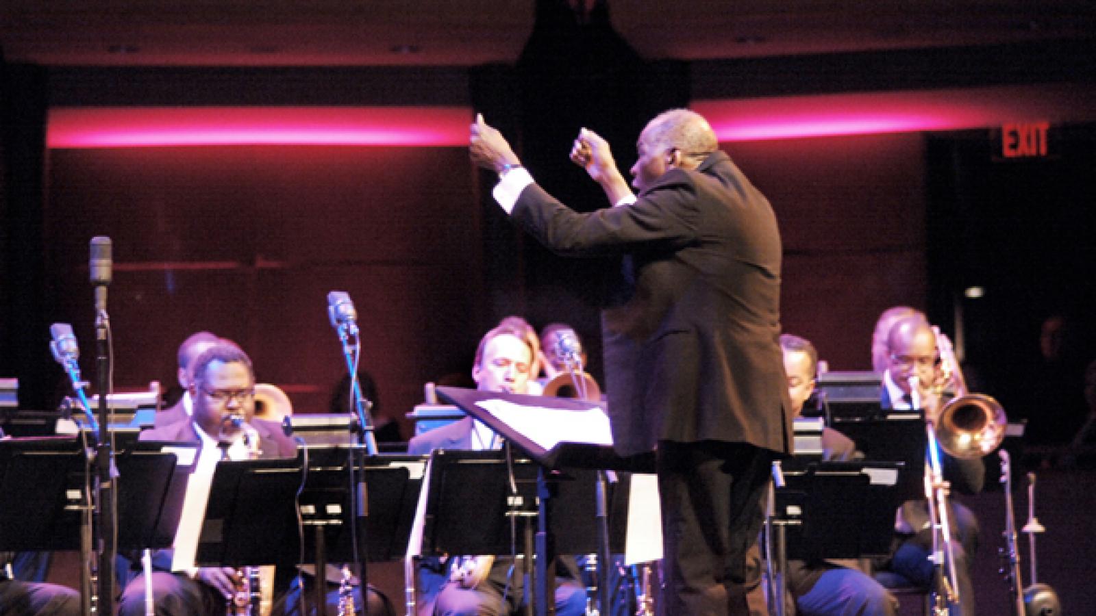 A man stands facing an orchestra with his arms raised