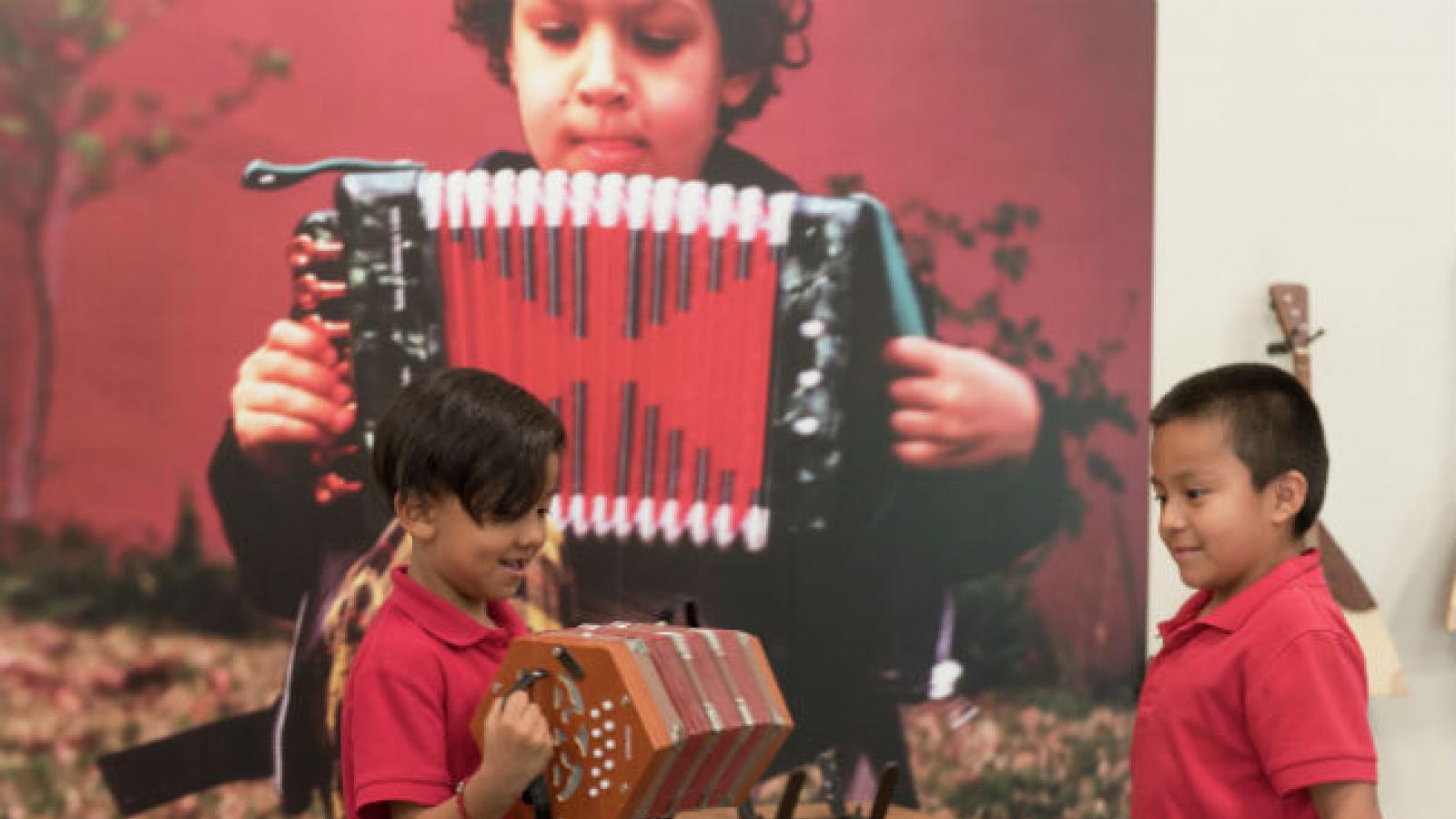 Two young boys play an accordion in front of a large picture of someone playing an accordion