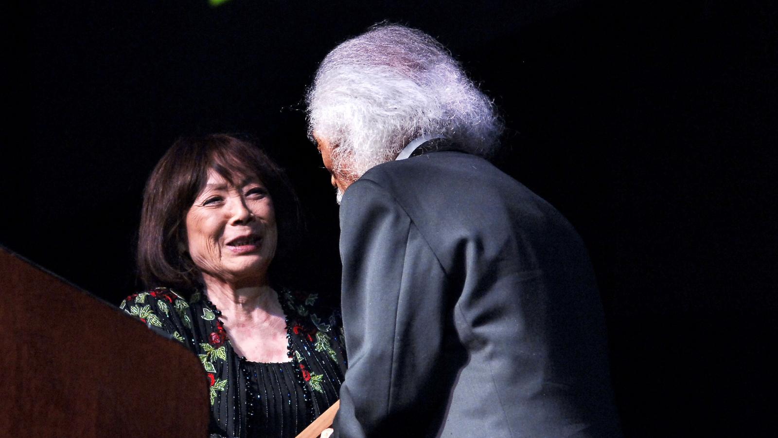 Japanese woman shaking hands with white-haired man in suit on stage. 
