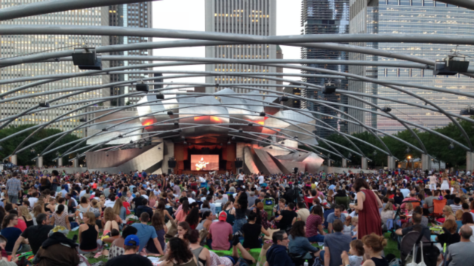 The silver Pritzker Pavilion in with audience in front sitting on grass