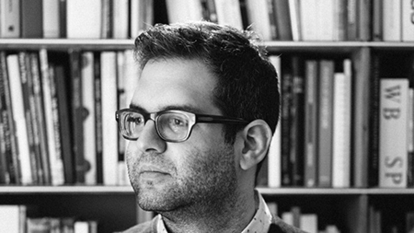 Portrait of man wearing dark glass frames in front of a bookcase. 