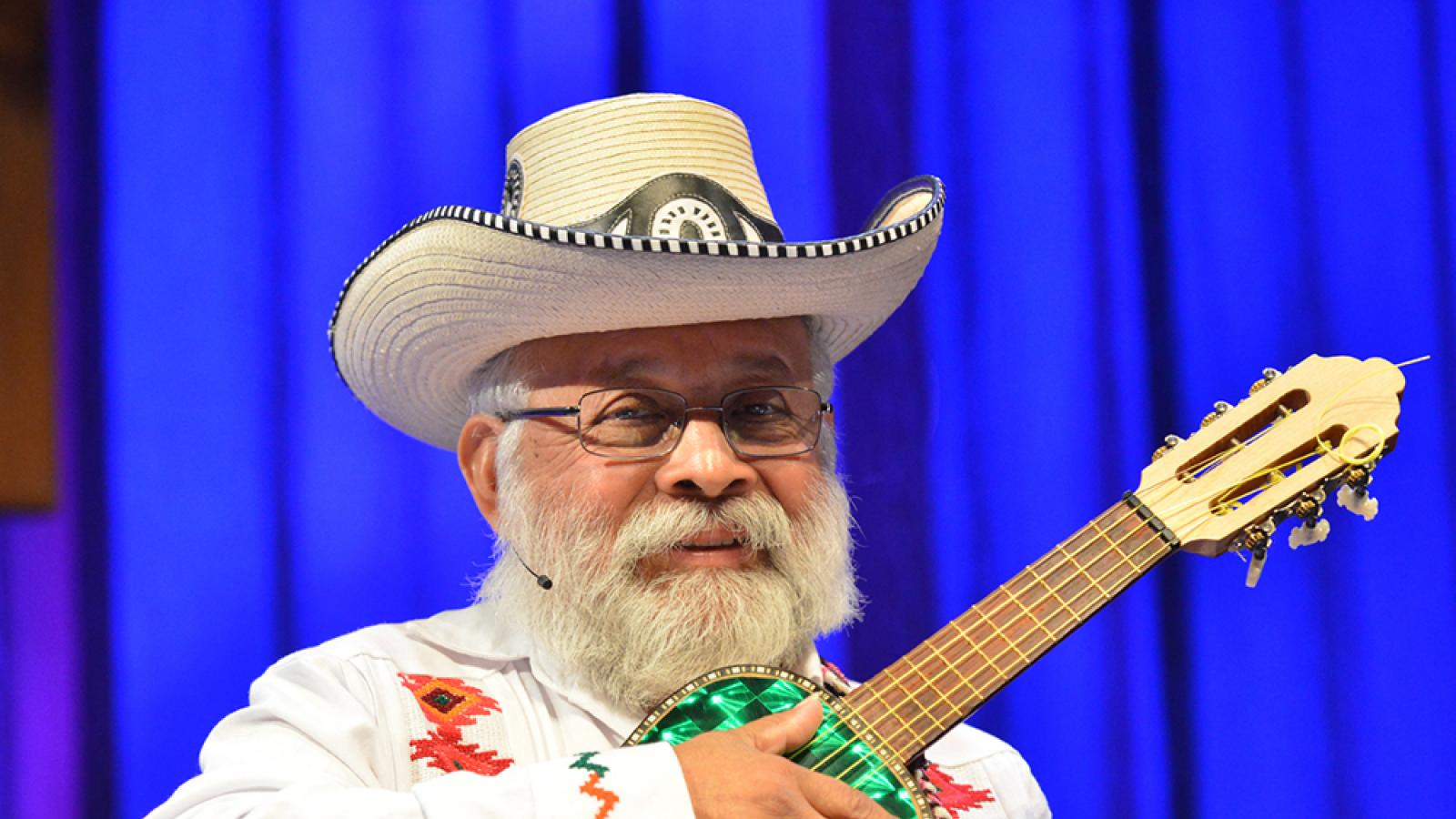 Musician with white beard in white suit and hat holding small guitar.