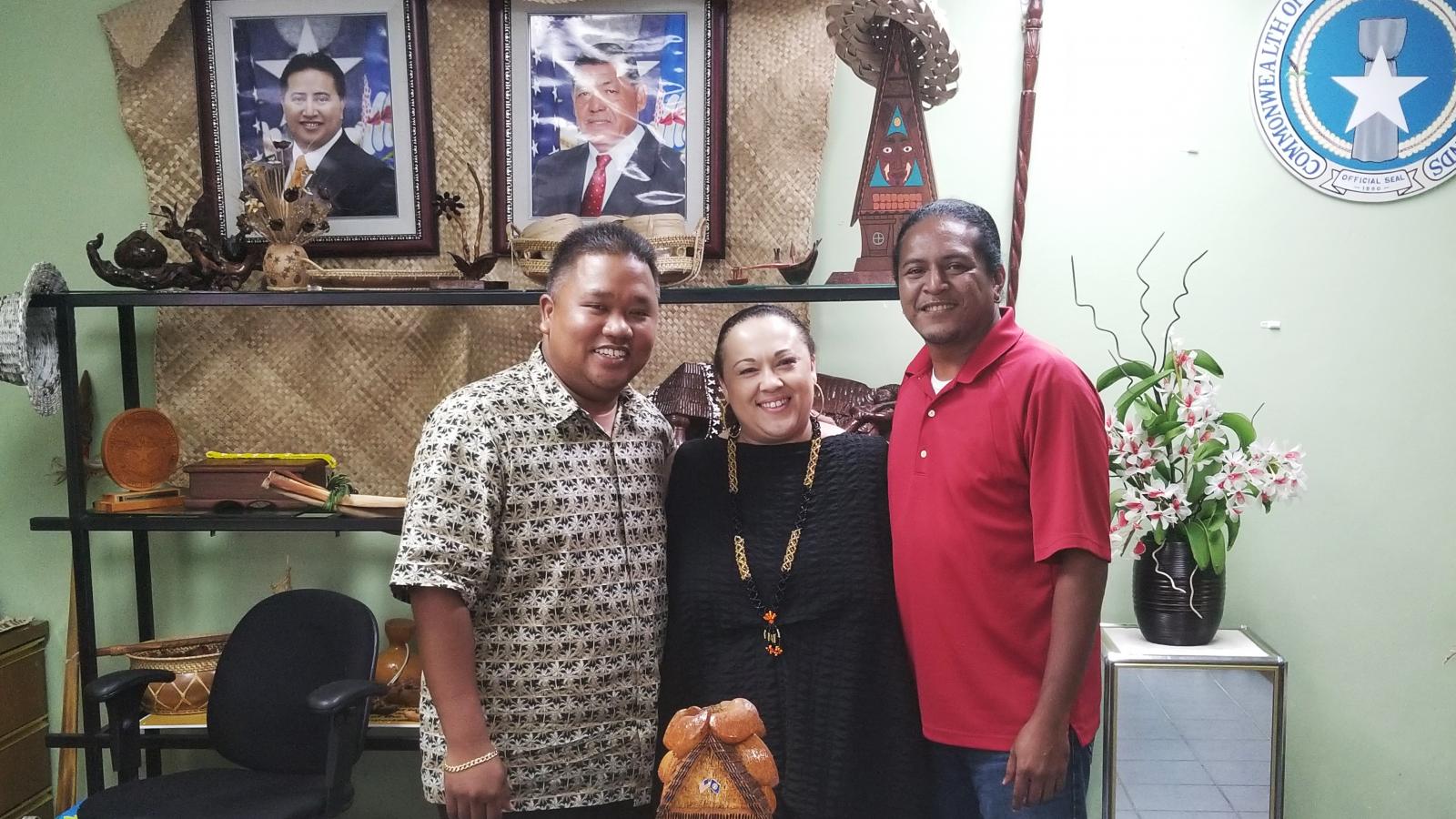Pam Breaux with staff from the Commonwealth Council for Arts and Culture A woman stands between two men amidst cultural objects from the Northern Mariana Islands