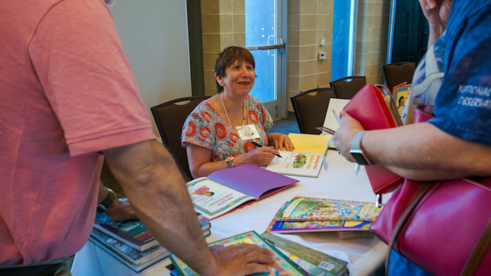 a woman sits at a conference table covered with children's book signing a copy of a book while a couple of adults look on