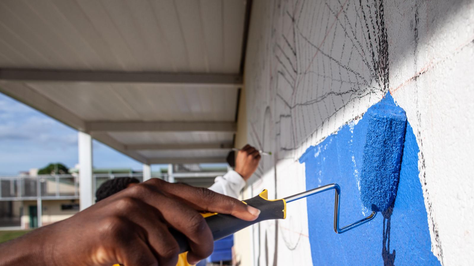 A hand in the foreground rolls blue paint on a cement block wall in the creation of a mural