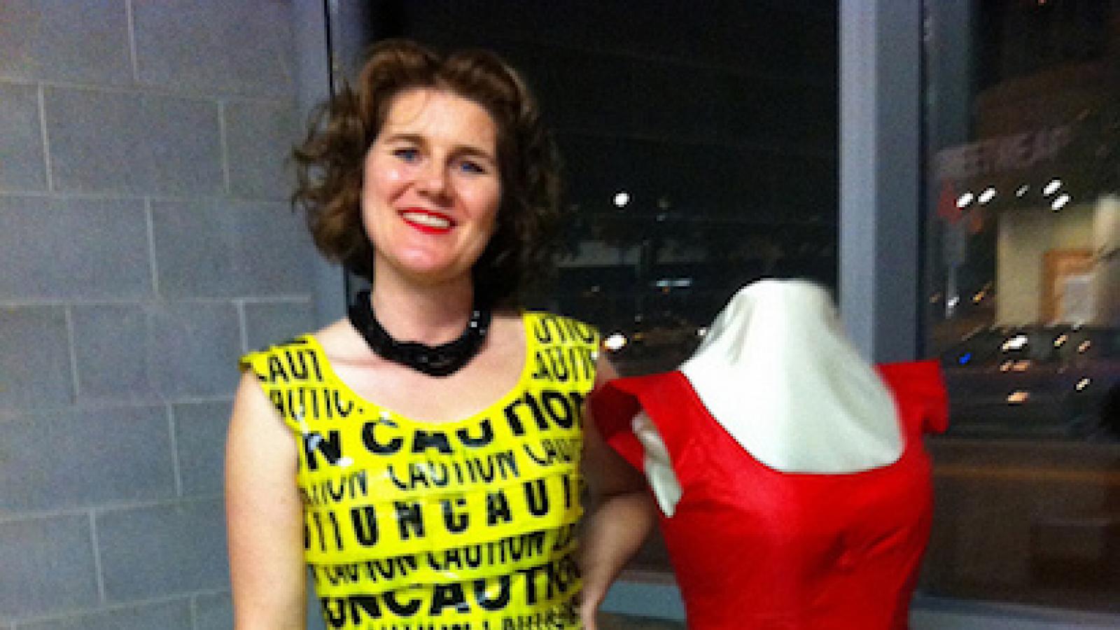 a woman wears a fitted dress made of caution tape and stands next to a dressmakers model wearing a red dress with a scalloped hi lo bottom