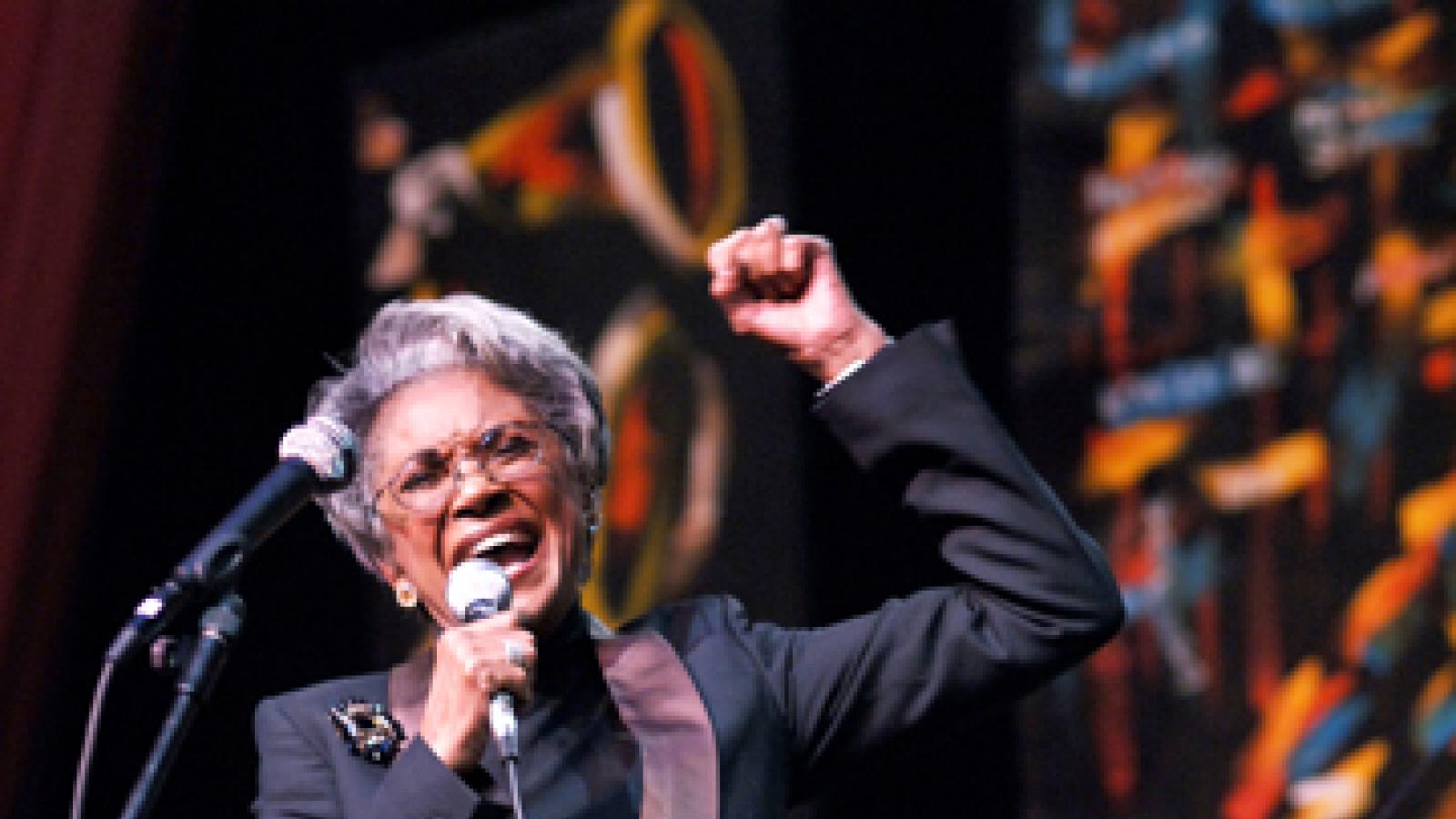 A woman in a black suit holds a microphone singing on a stage.