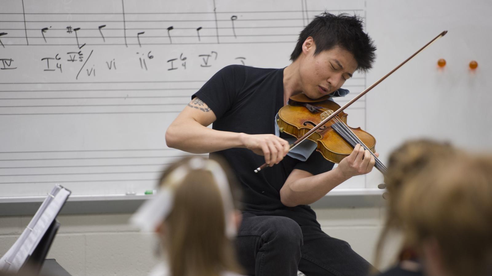 A man performs on the violin in a classroom while students watch.
