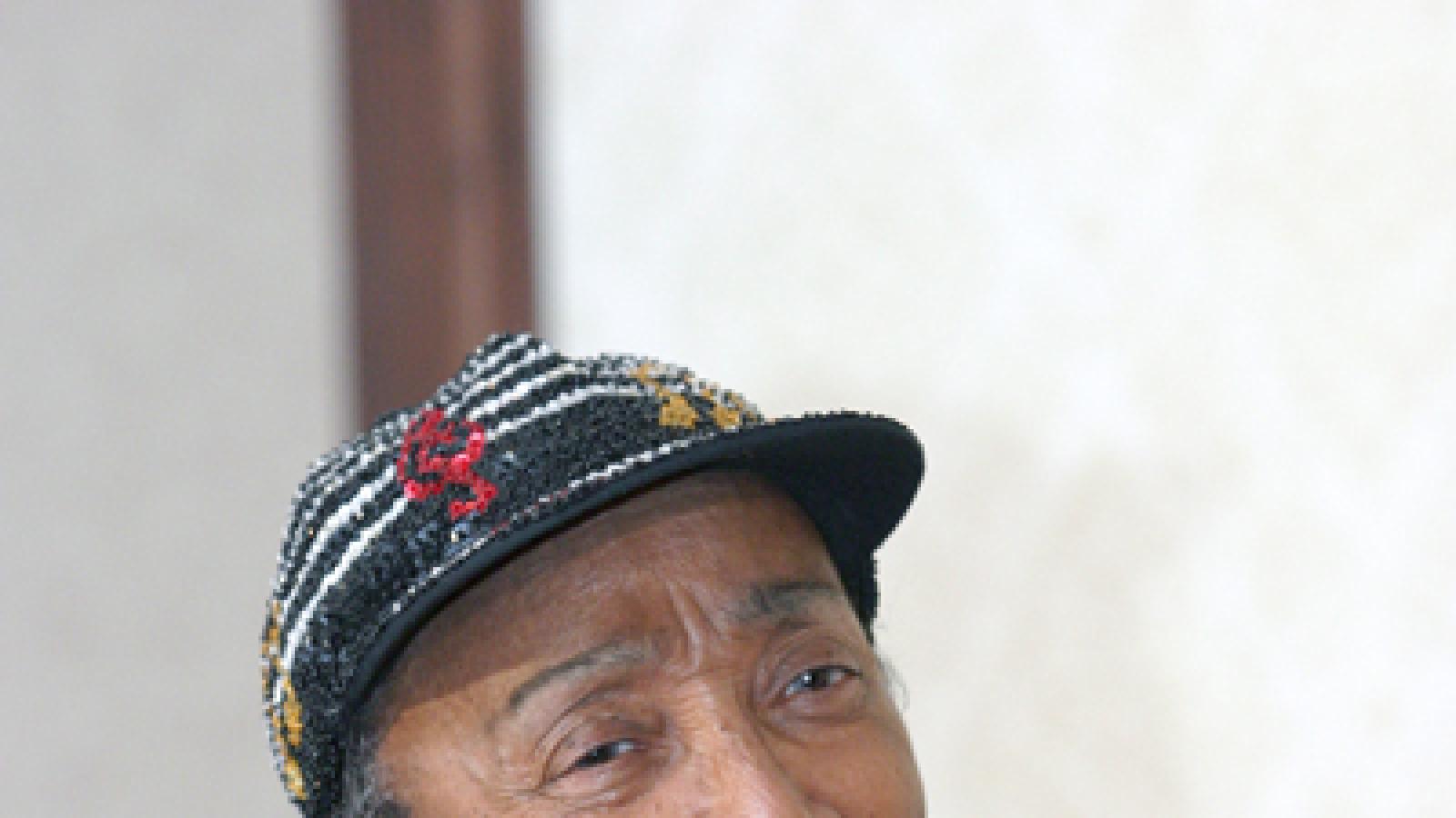 A woman wearing a red shirt and brightly sequined hat and vest speaks into a microphone.