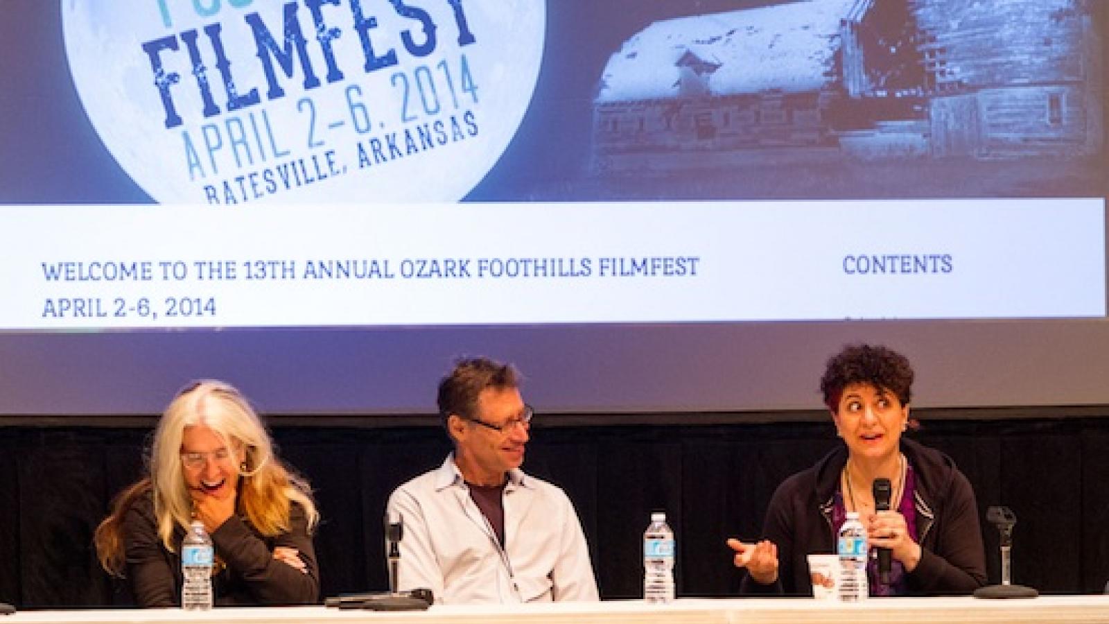 A woman, a man, and another woman holding a microphone and answering a question sit behind a long table with a film screen behind them