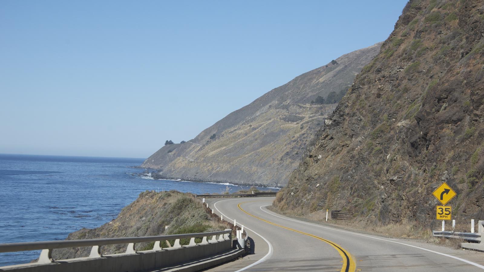 Empty road with the Pacific ocean to the left and mountains to the right.
