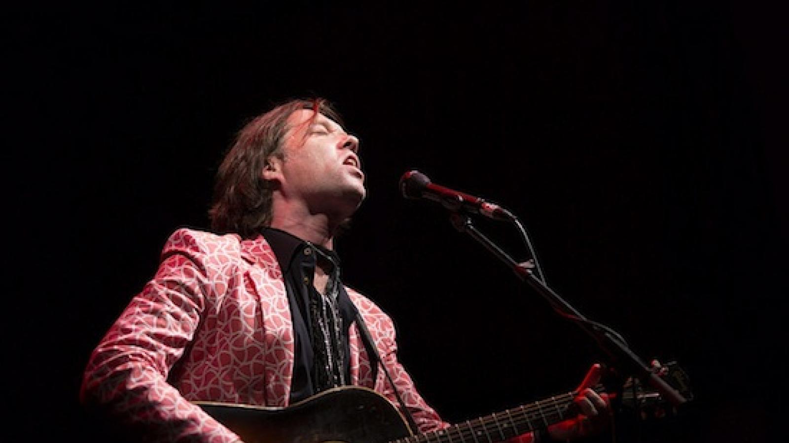 a white man with medium long hair in a patterned red jacket plays a guitar and sings into a microphone