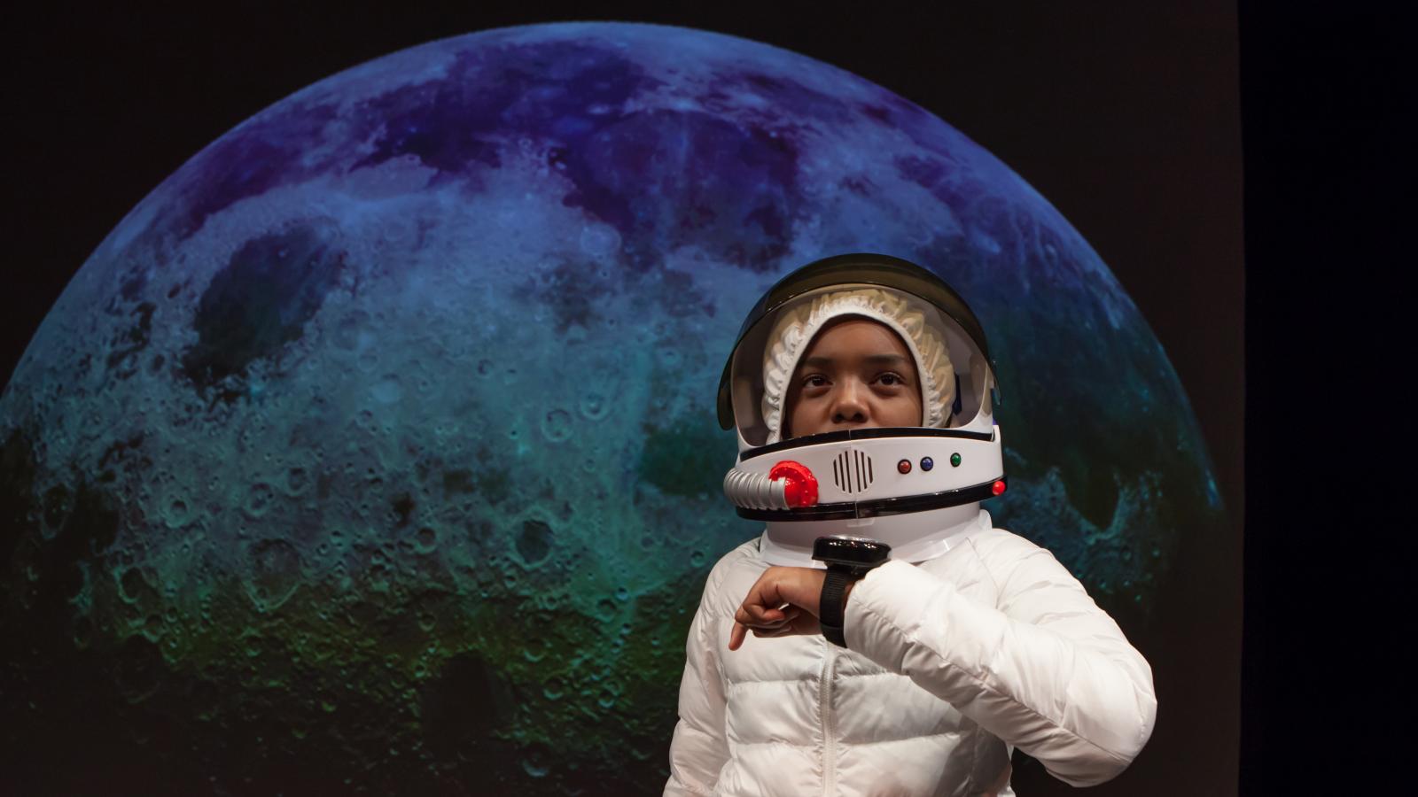 Boy in astronaut suit in front of photo of the moon. 